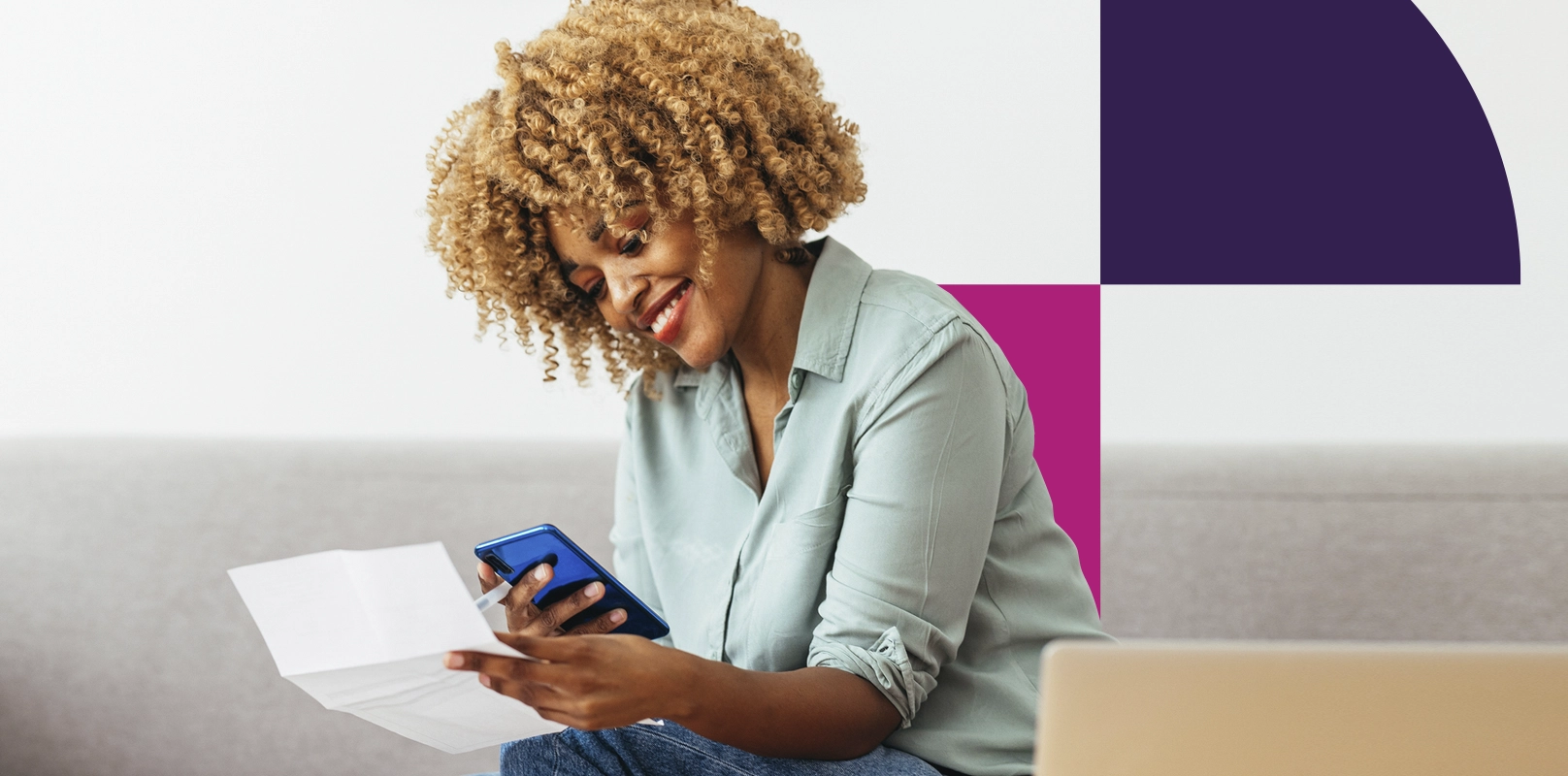 Smiling woman is sitting on a sofa and holding a document while looking at a smartphone in her hand.